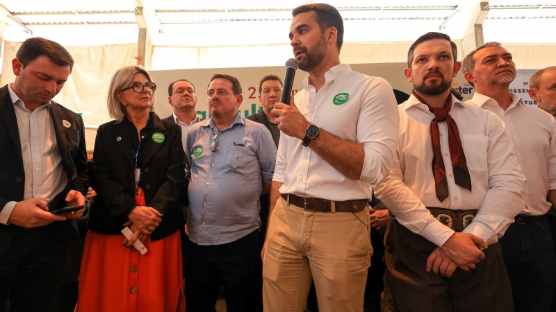 Num ambiente fechado, várias pessoas estão em pé, ao redor de Leite, também em pé, que fala ao microfone. Ao fundo, há um painel de cor branca, cujos escritos estão tapados pelas pessoas.