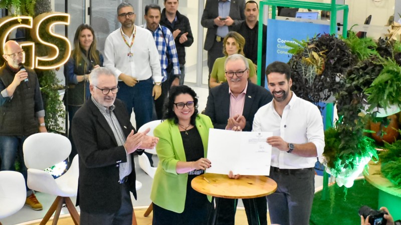 A imagem mostra, em primeiro plano, cinco pessoas em torno de uma mesa redonda de madeira clara. Quatro delas &mdash; tr&ecirc;s homens e uma mulher &mdash; est&atilde;o de p&eacute; e aplaudindo, enquanto um quinto homem, de camisa social branca, segura um documento com as duas m&atilde;os. H&aacute; um p&uacute;blico e outros indiv&iacute;duos ao fundo, em um cen&aacute;rio que se assemelha a um evento. No canto superior esquerdo, um letreiro luminoso exibe a sigla "GS".