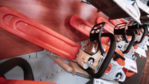 Foto de uma prateleira de loja com v&aacute;rias motosserras da marca STIHL expostas lado a lado. Em primeiro plano aparece uma motosserra branca, laranja e preta, identificada como modelo MS 261, com o sabre protegido por uma capa laranja. Ao fundo, outras motosserras semelhantes est&atilde;o alinhadas, com etiquetas de pre&ccedil;o e informa&ccedil;&otilde;es sobre os produtos. A parede do expositor &eacute; vermelha, com o nome STIHL em destaque.