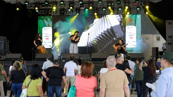 Fotografia de um show musical em um palco iluminado por luzes amarelas e verdes. Ao centro, um m&uacute;sico toca acorde&atilde;o; &agrave; esquerda, outro toca um tambor, e &agrave; direita, um terceiro m&uacute;sico toca viol&atilde;o sentado em um banco. Ao fundo, um grande painel de LED exibe uma imagem ampliada de um acorde&atilde;o. Na frente do palco, h&aacute; uma plateia numerosa, de costas para a c&acirc;mera, acompanhando a apresenta&ccedil;&atilde;o. A estrutura met&aacute;lica do palco est&aacute; repleta de refletores e equipamentos de ilumina&ccedil;&atilde;o, e h&aacute; fuma&ccedil;a c&ecirc;nica espalhada pelo ambiente.