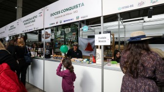 Foto mostra bancas num corredor do Pavilh&atilde;o da Agricultura Familiar. Numa delas, ao fundo, se v&ecirc; a bandeira do Estado de Minas Gerais, com o fundo branco e um tri&acirc;ngulo vermelho no centro. H&aacute; uma menina na frente da banca com um bal&atilde;o verde. Outras mulheres est&atilde;o visitando outras bancas. 