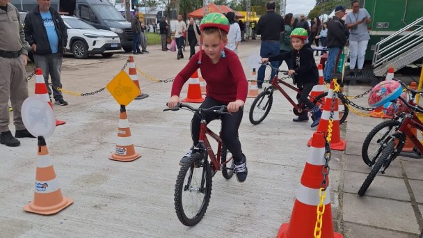 Foto de uma atividade educativa de tr&acirc;nsito realizada ao ar livre. Em primeiro plano, uma crian&ccedil;a usando capacete verde pedala uma bicicleta por um circuito sinalizado com cones laranja, correntes e placas. Ao fundo, outra crian&ccedil;a tamb&eacute;m participa da atividade com uma bicicleta, enquanto adultos observam. H&aacute; ve&iacute;culos estacionados, estruturas de estandes e circula&ccedil;&atilde;o de pessoas no local. O c&eacute;u est&aacute; nublado.